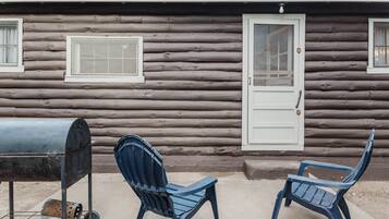 A log cabin with two blue plastic chairs, a grill, and a door.