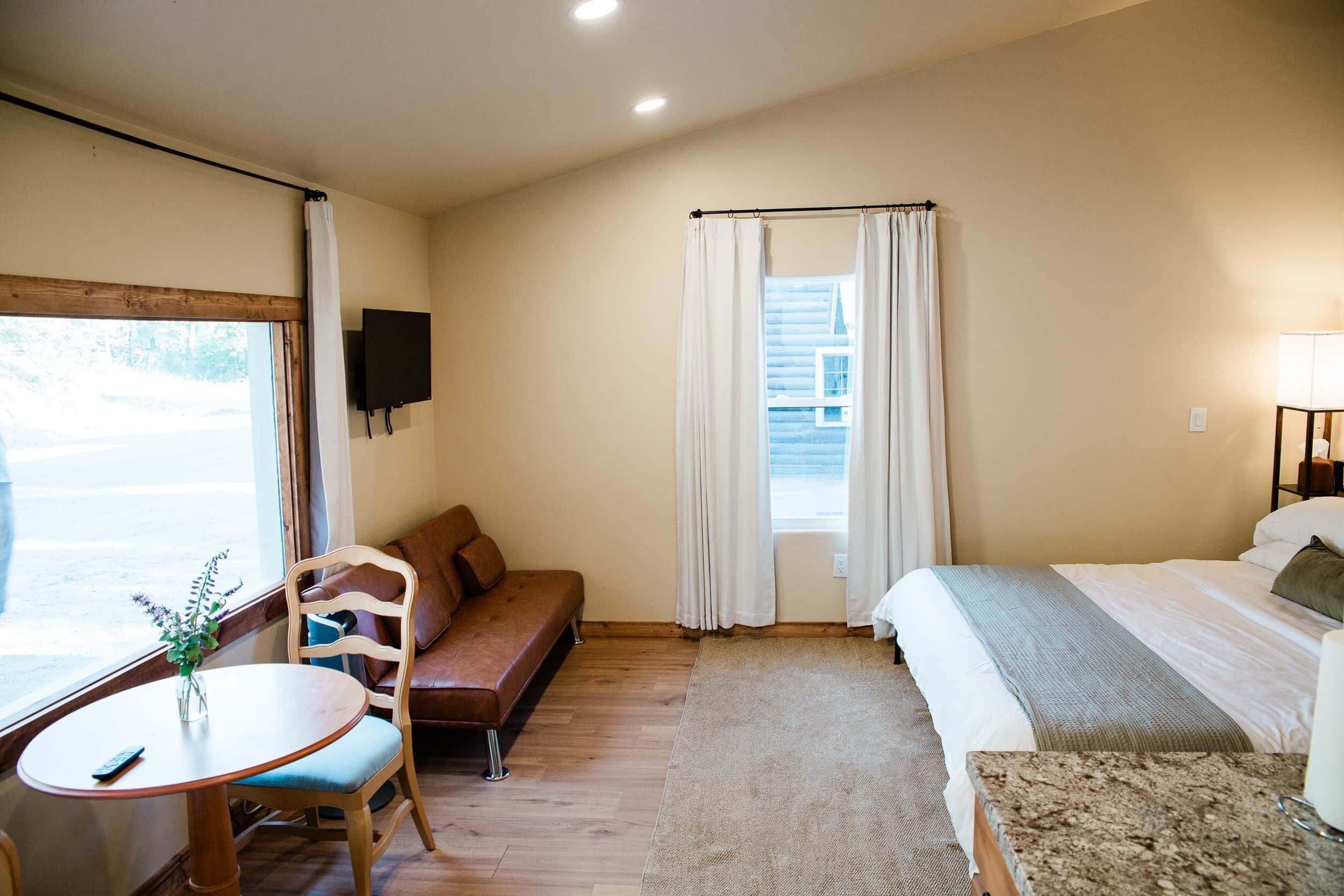 A hotel room with a bed, a sofa, a small table with a vase, a TV mounted on the wall, and a window with curtains.