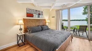 A bedroom with a large bed, two bedside lamps, a view of a lake through a glass door, and a painting on the wall.