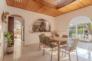 A dining area with a wooden table and patterned chairs, a kitchen with white cabinets, and a view of a balcony with a railing.