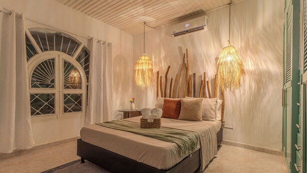 A bedroom with a bed, two hanging lamps, a window with curtains, and a wall-mounted air conditioner.