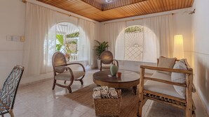 A room with a wooden ceiling, a round table, two chairs, a sofa, and a small table with a vase and cup.