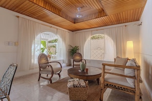 A room with a wooden ceiling, a round table, two chairs, a sofa, and a small table with a vase and cup.
