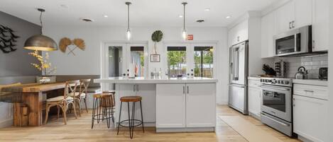 Une cuisine moderne avec des meubles blancs, des appareils en acier inoxydable, un coin repas en bois et des luminaires suspendus.