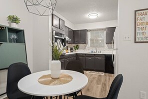 A modern kitchen with dark cabinets, a white countertop, and a dining area with a white table and chairs. - Modern Apartment near The Domain (Austin)