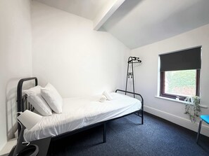 A single bed with a metal frame, white bedding, a small bedside table, a window with a view of greenery, and a ceiling with a skylight. - Comfortable Single Room in Wigan House Share (Wigan)