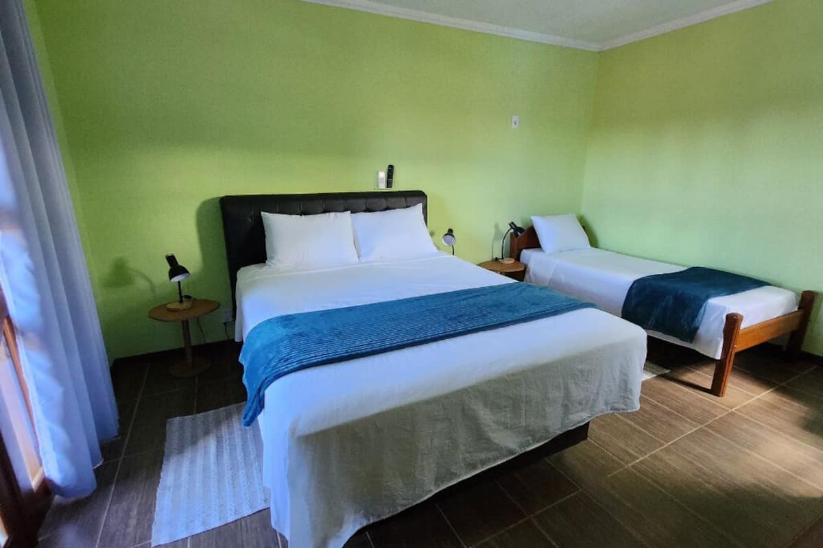 Habitación de hotel con una cama grande, una cama más pequeña, una mesita de noche, una lámpara y una ventana con cortinas.