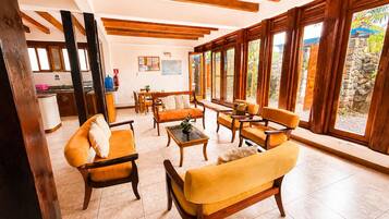 A spacious living area with wooden furniture, large windows, and a kitchen in the background.