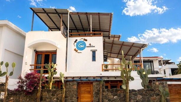 A modern two-story building with a stone wall, a wooden door, and a sign with a logo and text. - Modern, Spacious House with a Cozy Atmosphere and an Excellent Location (Puerto Ayora)