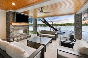 A modern outdoor living area with stone walls, a flat-screen TV, a fireplace, and a view of a lake with a staircase leading to an upper level. - Willow Landing w/Optional Boat Rental, Golf Simulator & Heated Pool (Troutman)