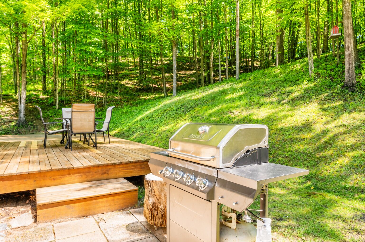 Una terraza de madera con una parrilla, sillas y una mesa en un área boscosa.