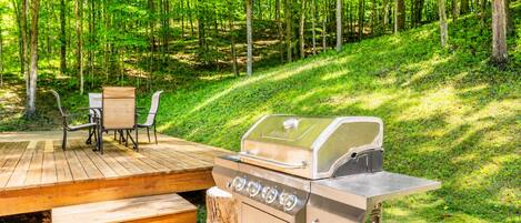 Una terraza de madera con una parrilla, sillas y una mesa en un área boscosa.