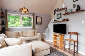Una sala de estar con sofá, reposapiés, mueble de televisión de madera y una silla.