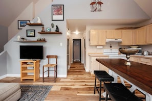 Una cocina moderna con armarios de madera, un comedor con mesa y banco de madera, un televisor sobre un mueble de madera y una escalera que sube a otro nivel.
