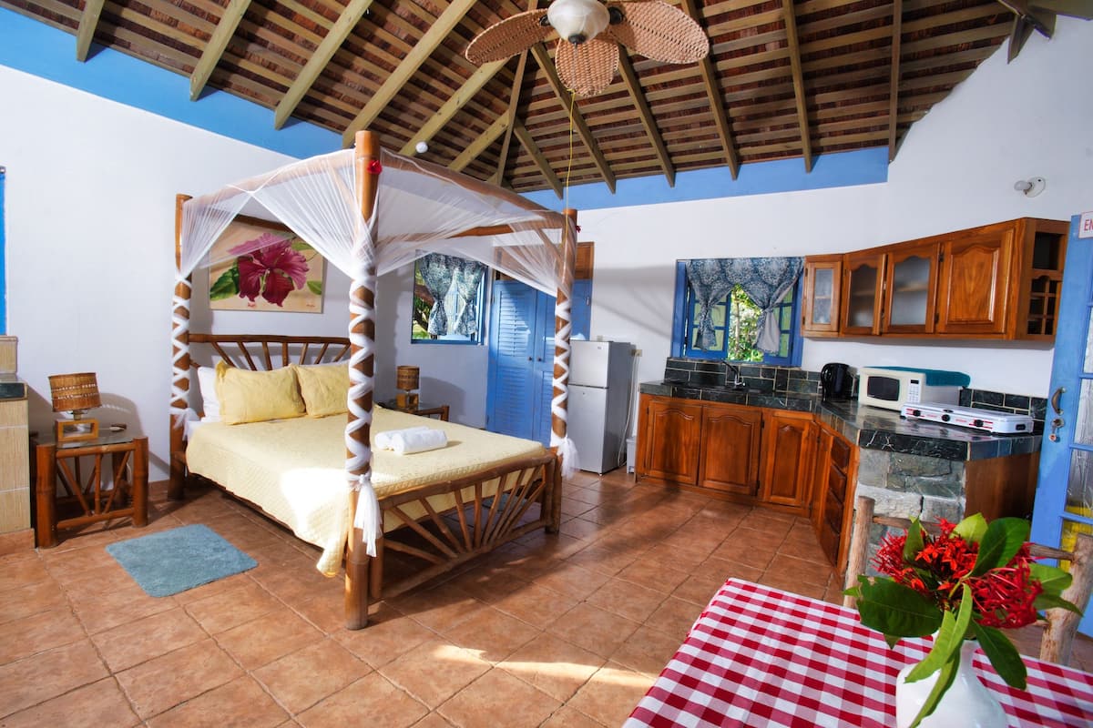 A bedroom with a four-poster bed, a small table with a vase, and a view of the kitchen and outside.