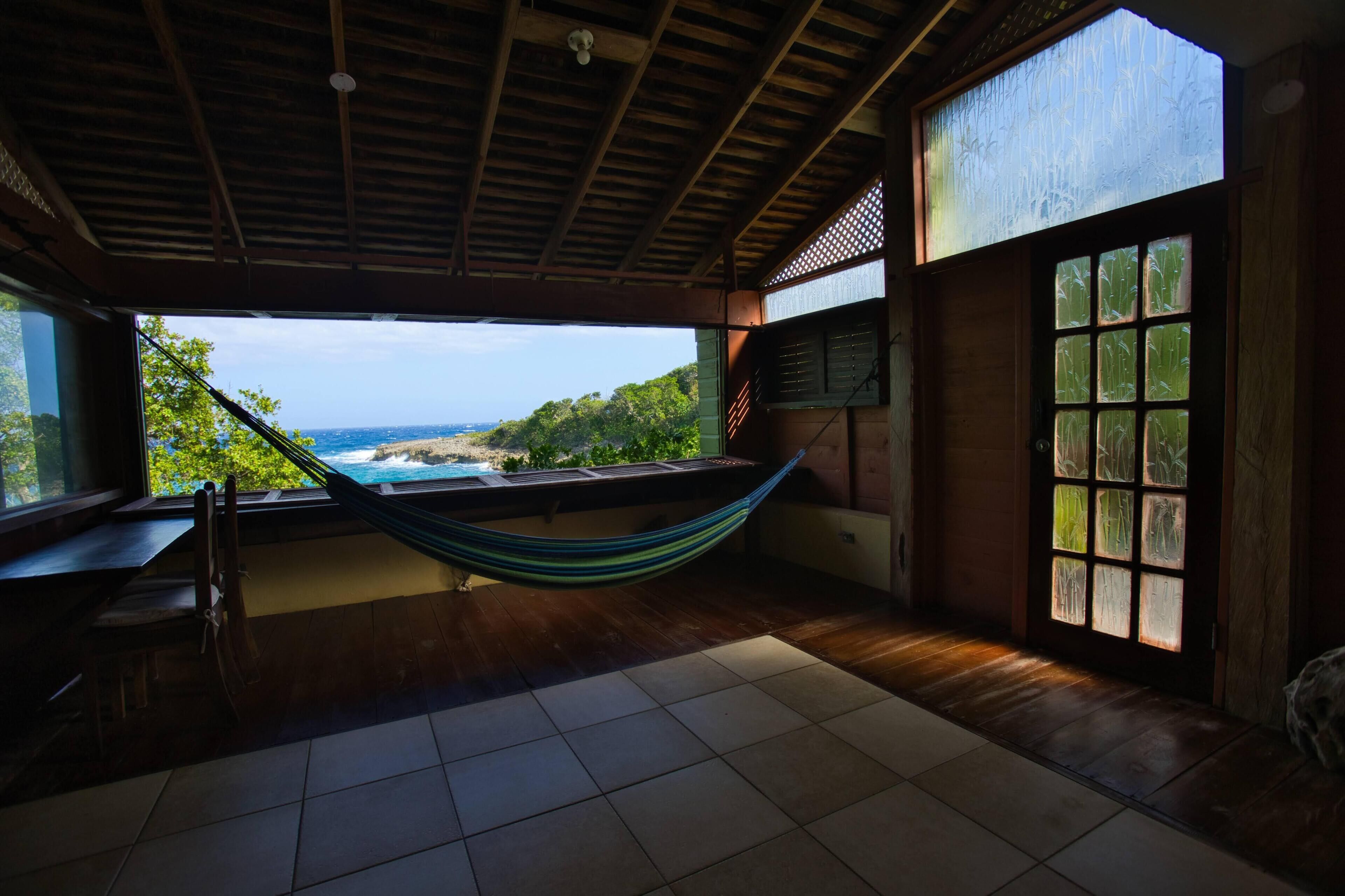A room with a hammock, wooden flooring, a view of the ocean, and a large window.