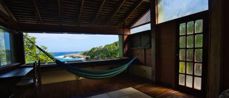 A room with a hammock, wooden flooring, a view of the ocean, and a large window.