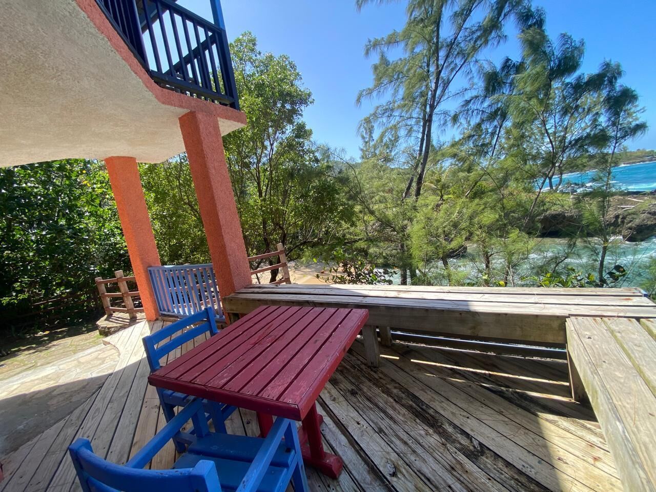 A wooden deck with a red table and blue chairs, overlooking a beach and ocean.