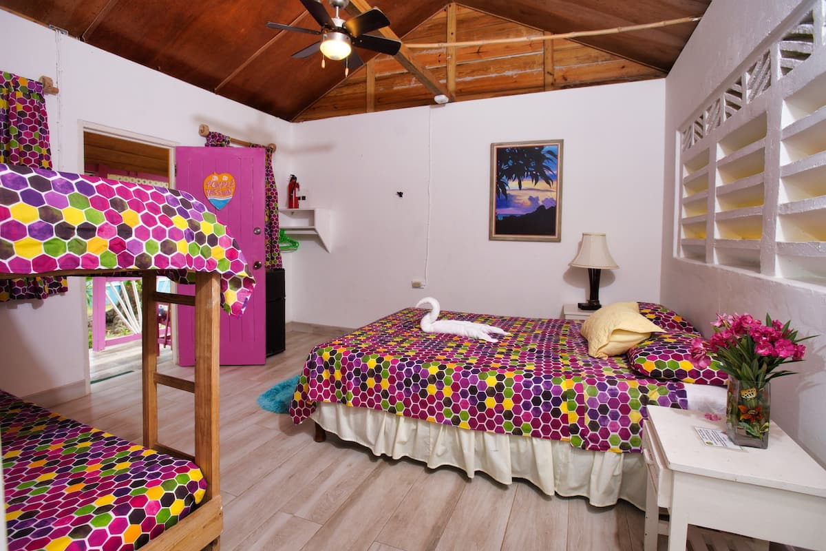 A bedroom with a bed, a vanity, a ceiling fan, a painting, and a window with a view.