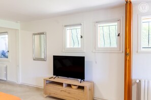 Ein Zimmer mit Fernseher auf einem Holzständer, zwei Fenstern mit Vorhängen und einer Heizung.