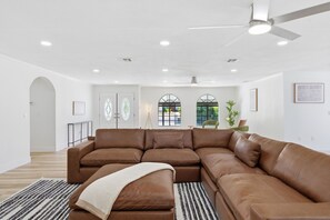 A spacious living room with a large brown sectional sofa, a ceiling fan, recessed lighting, and a view through a window.