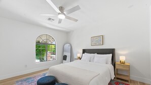 Une chambre à coucher avec un lit, un ventilateur de plafond, une fenêtre, un miroir, une table de chevet avec une lampe et un tapis coloré.