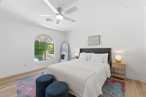 A bedroom with a bed, a ceiling fan, a window, a mirror, a bedside table with a lamp, and a colorful rug.