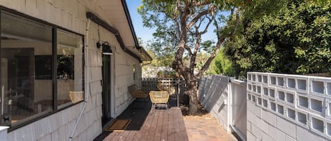 A covered outdoor area with a wooden deck, a white building with large windows, and a tree providing shade.