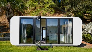 Un edificio moderno, blanco y modular con amplios ventanales y una puerta, rodeado de abundante vegetación.
