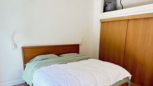 Ein Schlafzimmer mit einem Holzbettgestell, einer weißen Matratze und einer grünen Decke. An der Wand befinden sich ein Holzschrank und eine weiße Klimaanlage.