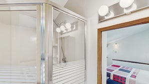 A shower with a glass door, a mirror with lights, and a bed with a quilt. - Cozy 3BR Condo - Fireplace - Tannersville (Pennsylvania)