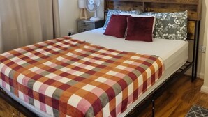 Un lit avec une couverture à carreaux, une tête de lit en bois, une table de chevet avec une lampe et une fenêtre avec des rideaux.