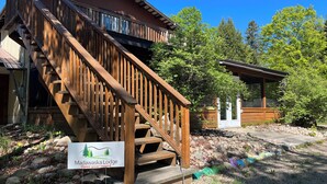 A wooden lodge with a staircase leading to a deck, surrounded by trees and a clear sky.