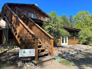 A wooden lodge with a staircase leading to a deck, surrounded by trees and a clear sky.