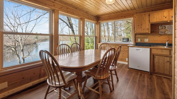 A wooden cabin with a dining table and chairs, large windows, a kitchen area, and a view of trees. - A 3 BR Lakefront Cabin on Balsam Lake w/Fire Pit and Sunset Views. Boat Rental (Birchwood)