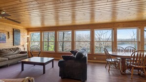 A spacious living room with a wooden ceiling, large windows, a sofa, a coffee table, and a dining area with chairs. - A 3 BR Lakefront Cabin on Balsam Lake w/Fire Pit and Sunset Views. Boat Rental (Birchwood)