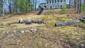 A modern house with a deck, surrounded by trees, and a fire pit area with chairs. - A 3 BR Lakefront Cabin on Balsam Lake w/Fire Pit and Sunset Views. Boat Rental (Birchwood)