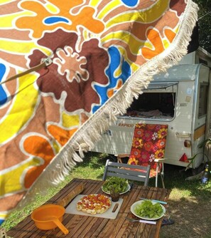 Una mesa con pizza, ensalada y una bebida, un mantel colorido, y una autocaravana al fondo.
