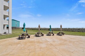 A patio area with picnic tables and umbrellas, a building with a blue exterior, and a grassy field in the background.