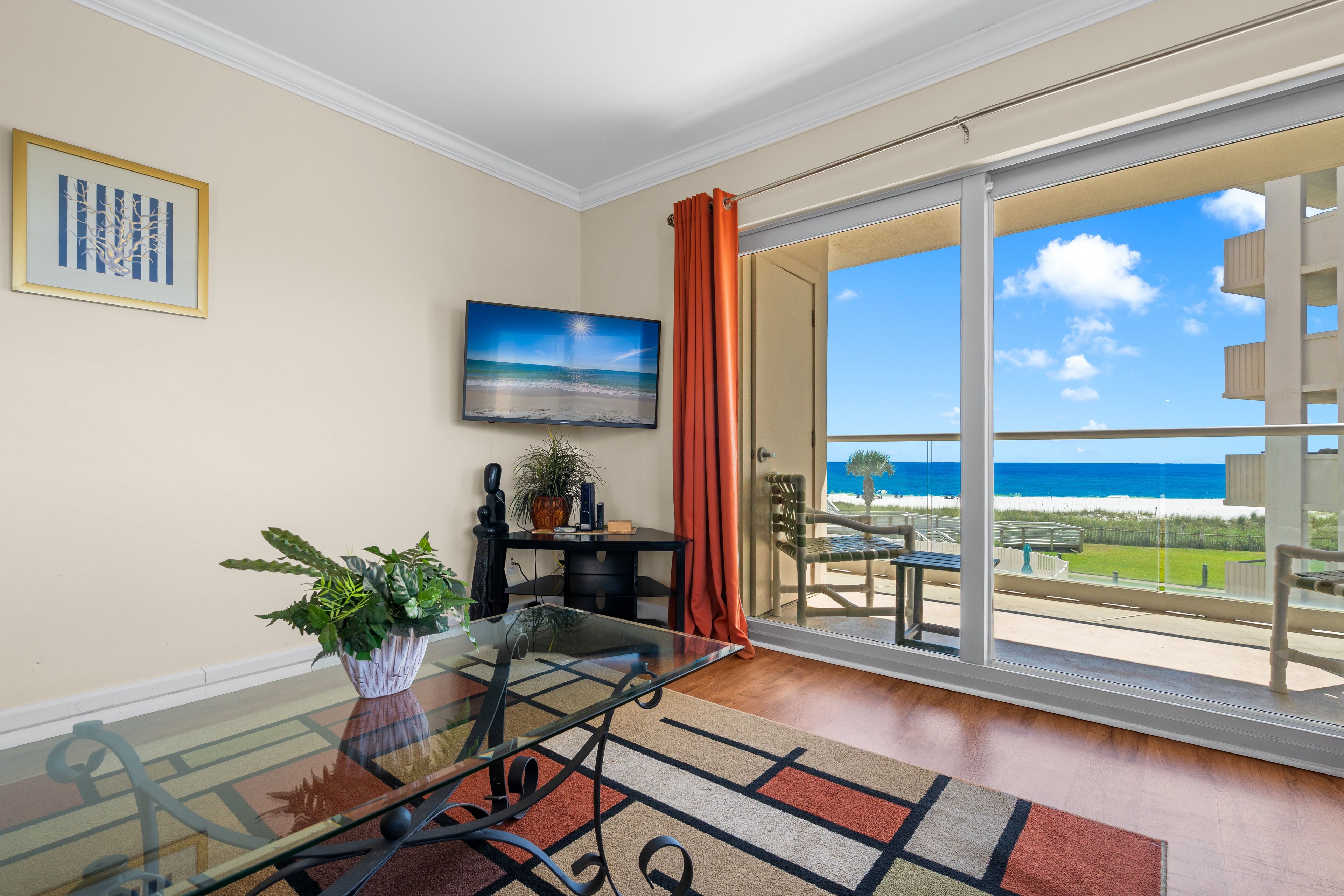 A room with a glass table, a potted plant, a painting, and a view of the ocean through large windows.
