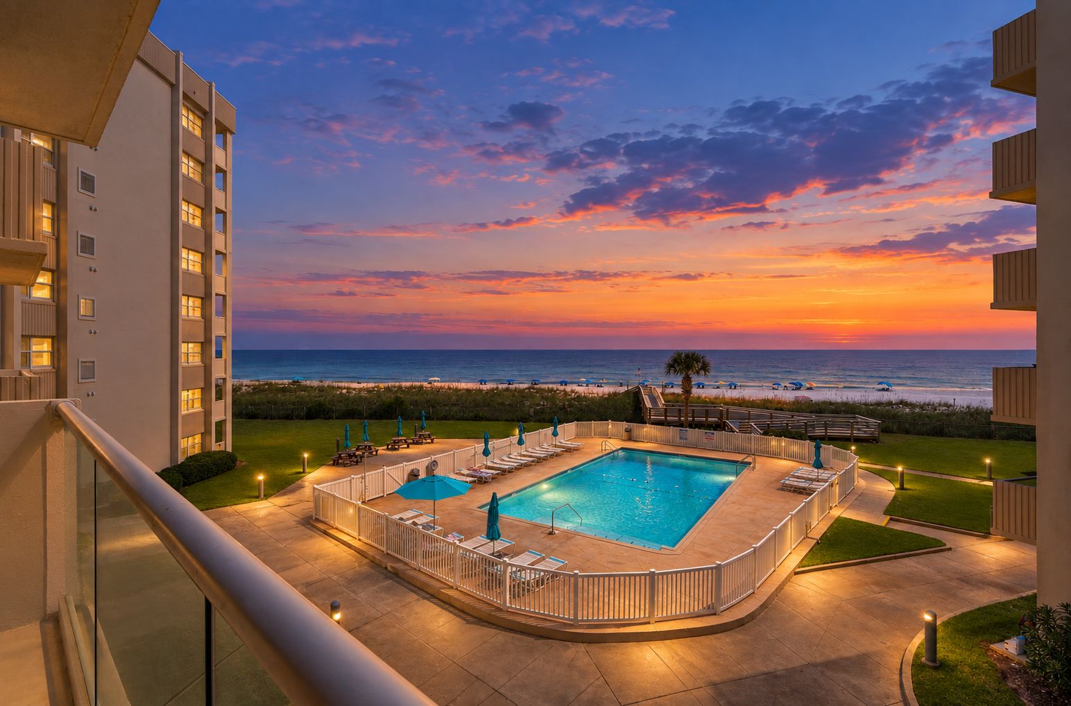 A pool area with a view of the ocean at sunset, surrounded by a fence and lounge chairs.