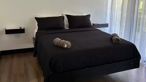 A modern bedroom with a large bed, two pillows, a bedside table, and a window with sheer curtains. - Duke's Place 2 (Quepos)