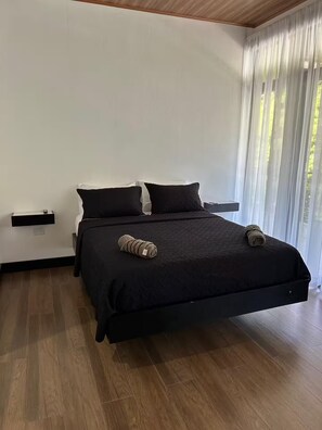 Ein modernes Schlafzimmer mit einem großen Bett, zwei Kissen, einem Nachttisch und einem Fenster mit leichtem Vorhang.