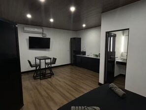 Ein modernes Zimmer mit Holzboden, einem Esstisch mit Stühlen, einem flachbildfernseher an der Wand, einer Küchenzeile mit Spüle und Arbeitsplatte sowie einem großen Spiegel.