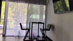 A room with a large window, a TV mounted on the wall, and a table with chairs. - Duke's Place 3 (Quepos)