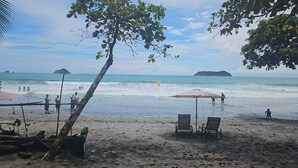 A beach with a few chairs and a table under a tree, a clear sky, and calm sea with distant islands. - Duke's Place 3 (Quepos)