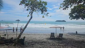 Ein Strand mit einigen Stühlen und einem Tisch unter einem Baum, ein klarer Himmel und ruhiges Meer mit fernen Inseln.