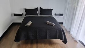 Una cama bien hecha con una colcha negra y dos almohadas, piso de madera y pared blanca con una mesita de noche pequeña.