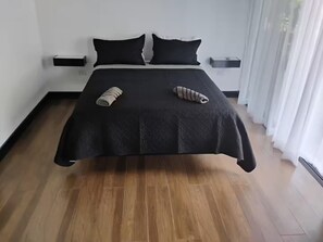 Ein ordentlich bezogenes Bett mit schwarzer Tagesdecke und zwei Kissen, Holzboden und weiße Wand mit einem kleinen Nachttisch.
