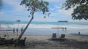 Ein Strand mit einigen Stühlen und einem Tisch unter einem Baum, ein klarer Himmel und ruhiges Meer mit fernen Inseln.